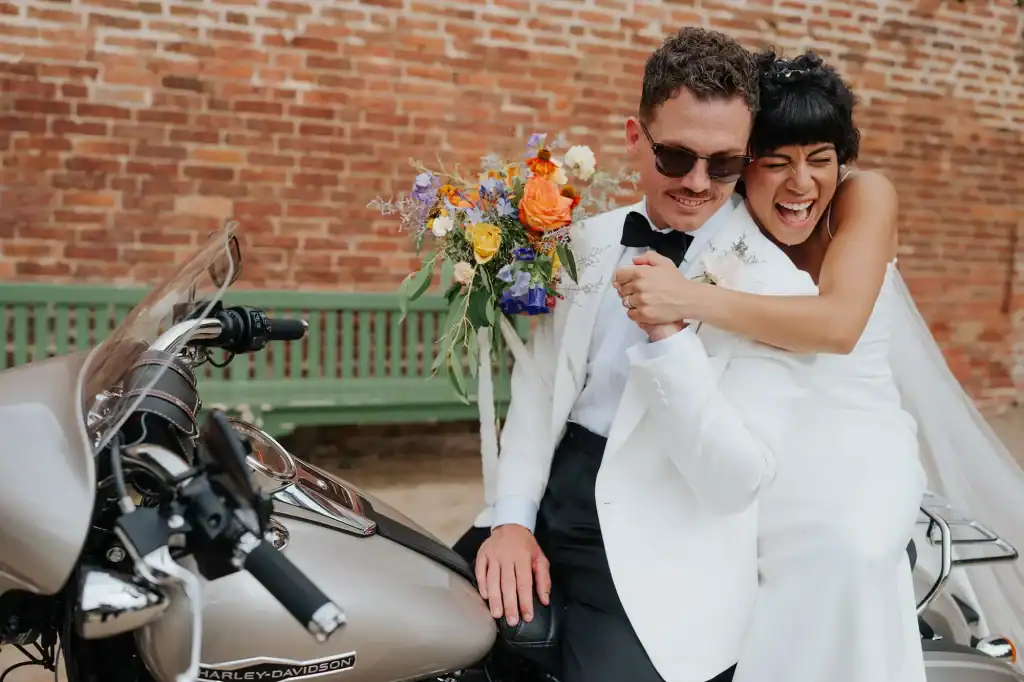 A bride and groom on a motorcycle holding a bouquet as she laughs whilst giving groom a big hug.