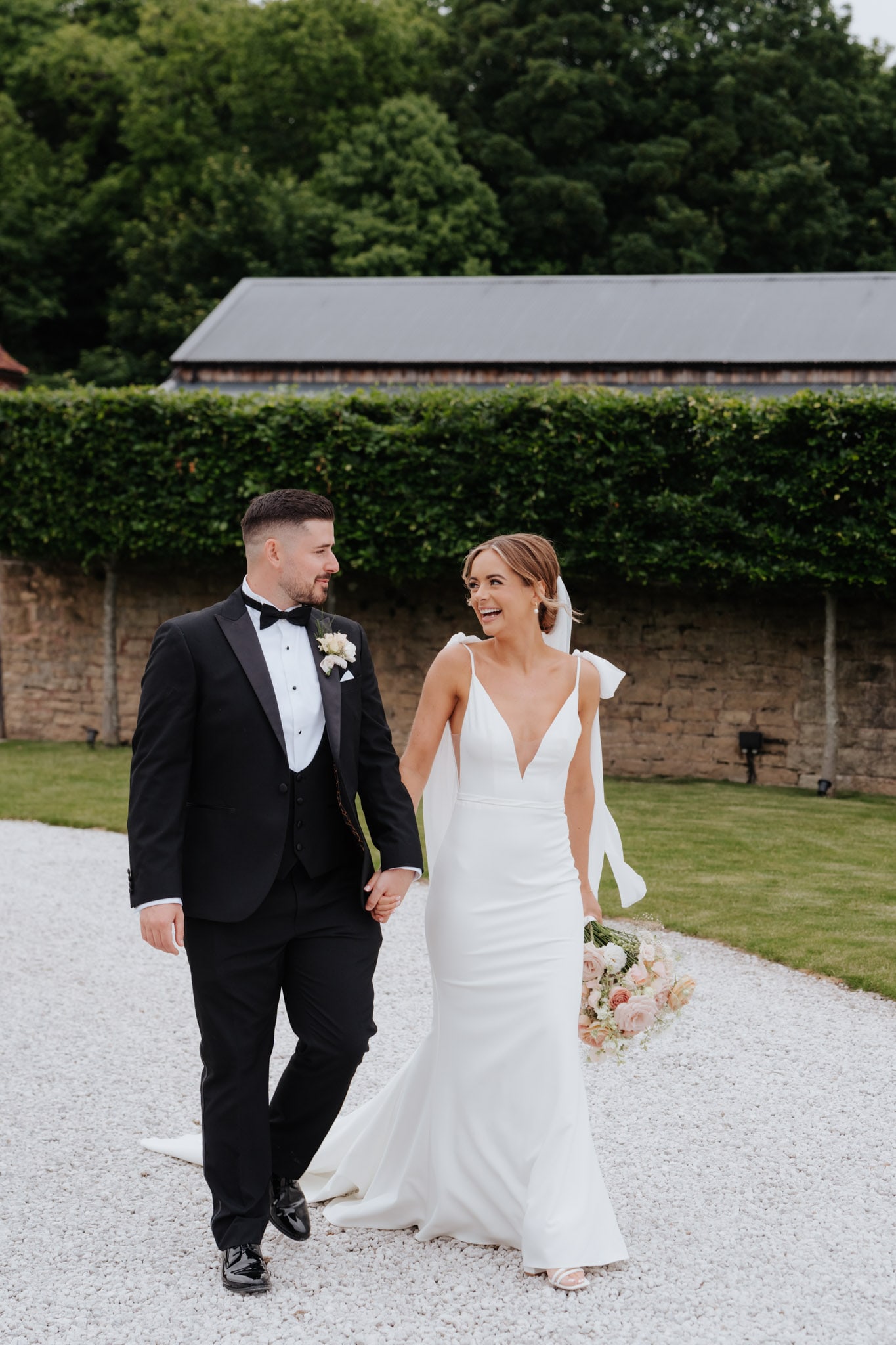 A bride and groom walk hand in hand outdoors. The bride wears a white dress with a deep neckline and bow sleeves, while the groom wears a black tuxedo. They are smiling at each other, with greenery and a stone wall in the background.