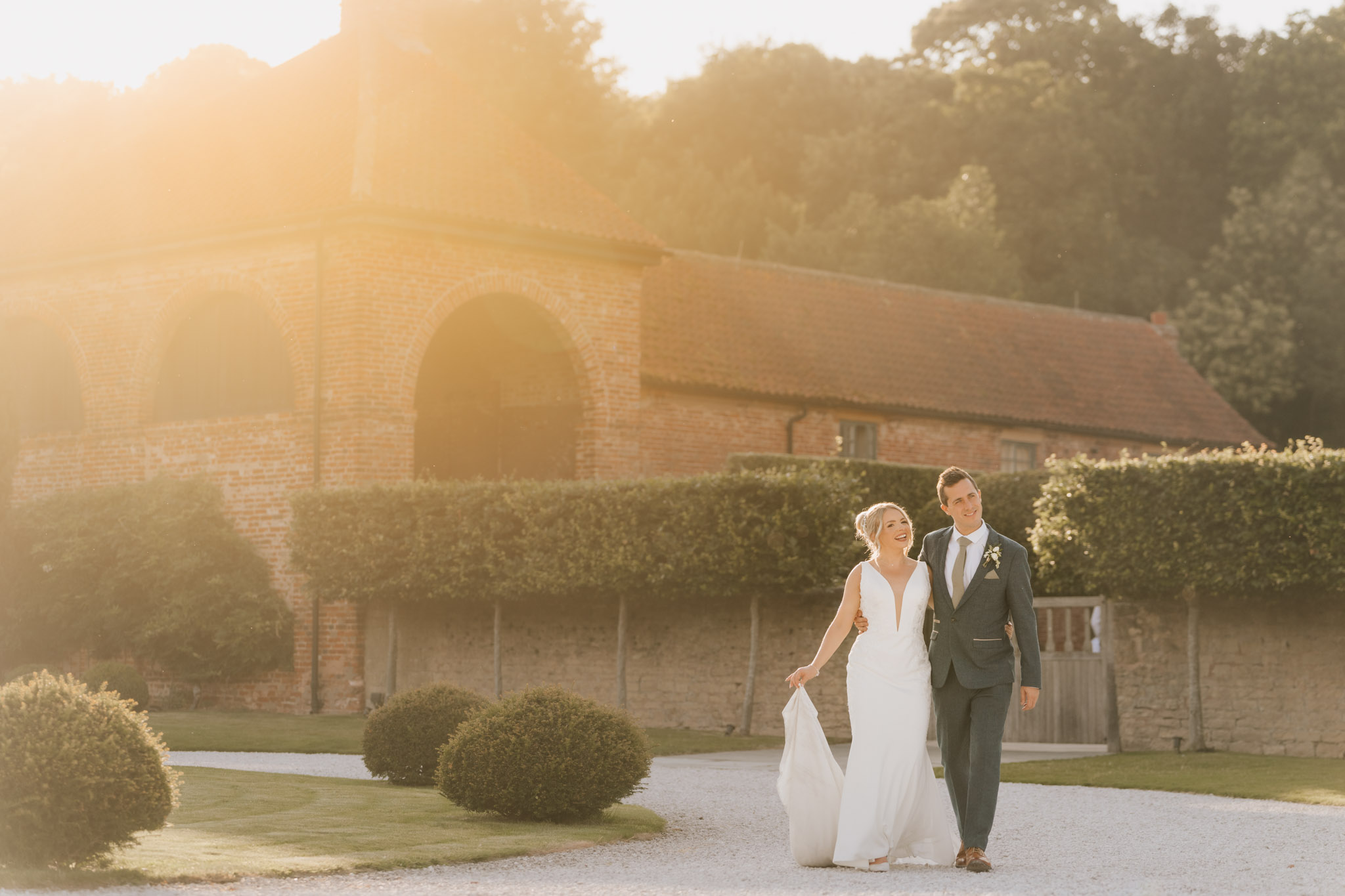 A bride and groom walk hand in hand on a gravel path, smiling, with Hazel Gap Barn’s rustic brick backdrop and lush greenery bathed in warm, golden sunlight.