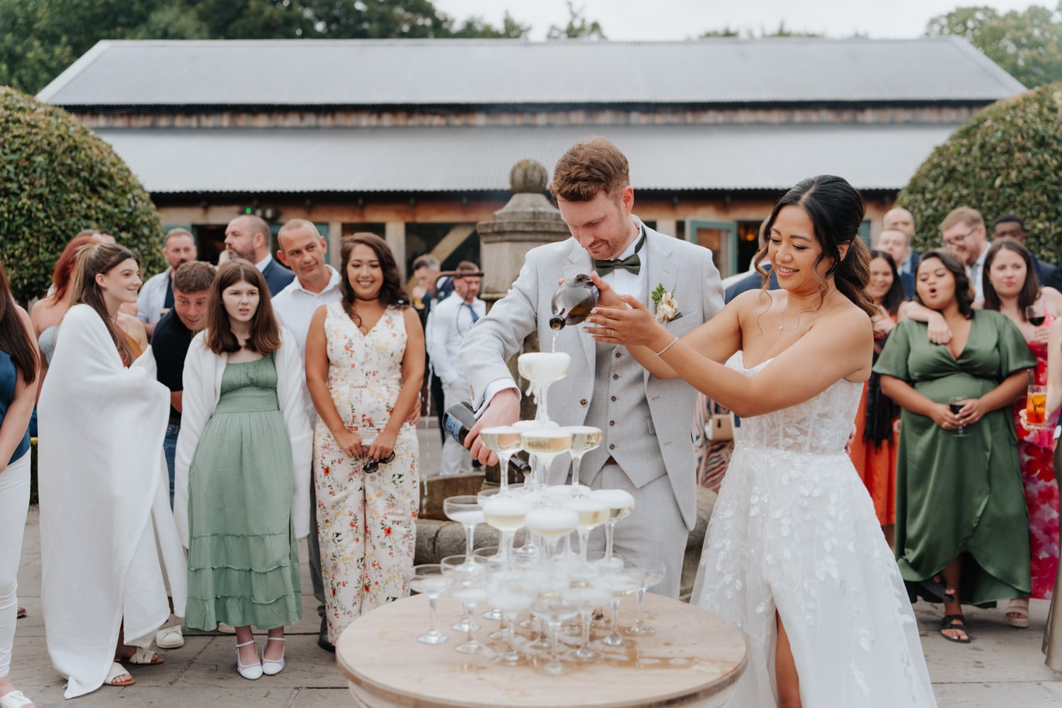 A bride and groom pour champagne into a pyramid of glasses during their outdoor wedding celebration, surrounded by smiling guests dressed in formal attire.