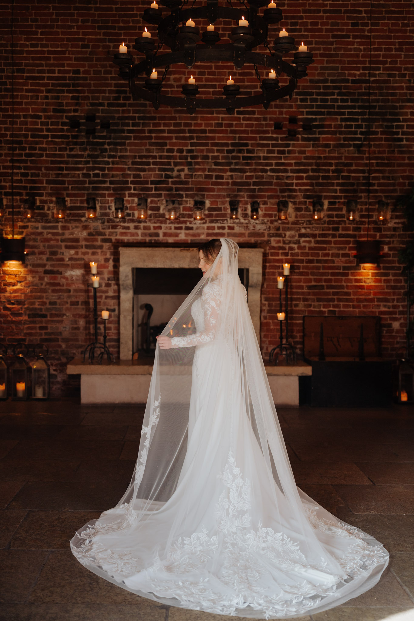 A bride in a long white gown with a lace train and veil stands in front of a brick fireplace, surrounded by lit candles. Chandeliers with candles hang above, creating a warm and elegant atmosphere.