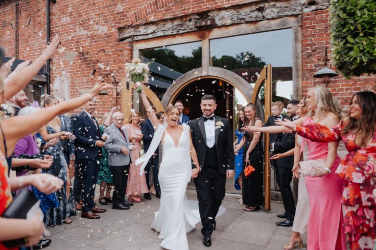 Bride points bouquet in the air whilst walking through confetti tunnel at Hazel Gap barn with her groom who is dressed in a black tuxedo