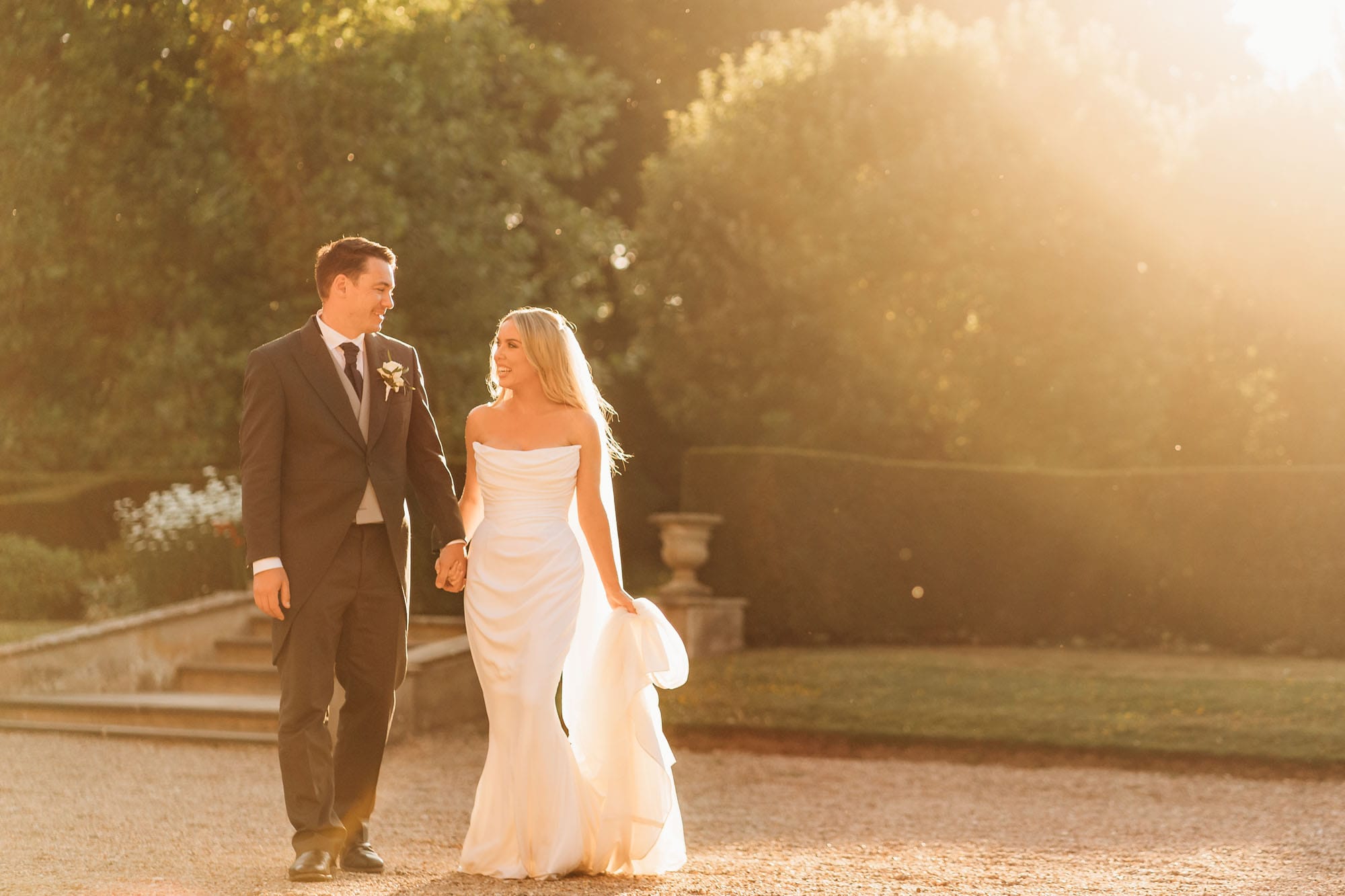 A bride in a white dress and a groom in a black suit walk hand in hand outdoors, bathed in warm sunlight, with greenery glowing behind them—perfectly captured by a Norwood Park Wedding Photographer.
