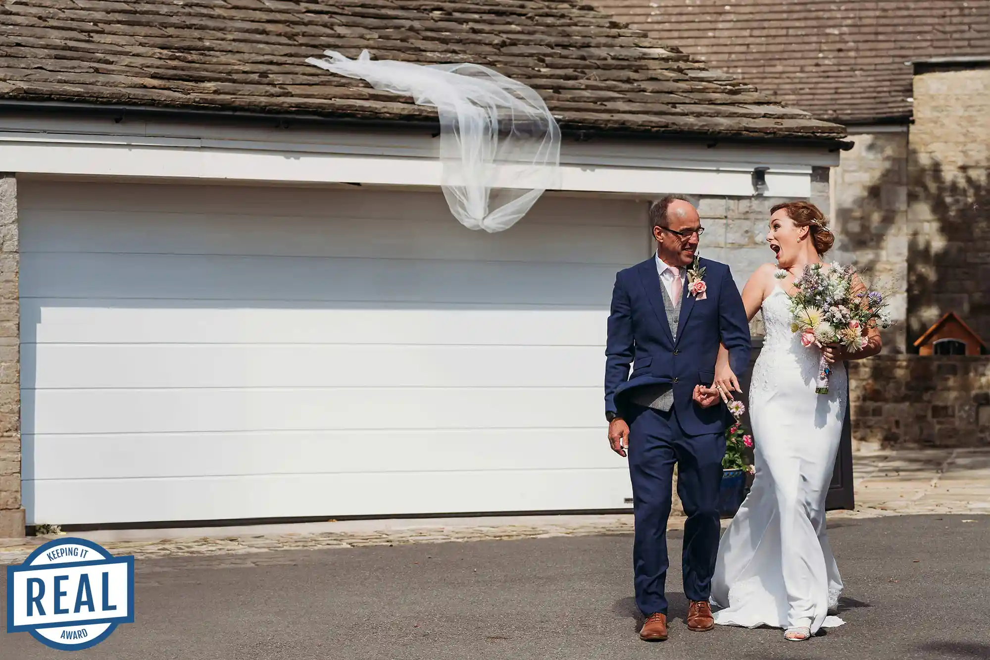Bride with a shocked look on her face, walking to her wedding arm in arm with dad because the wind causes her veil to fly off