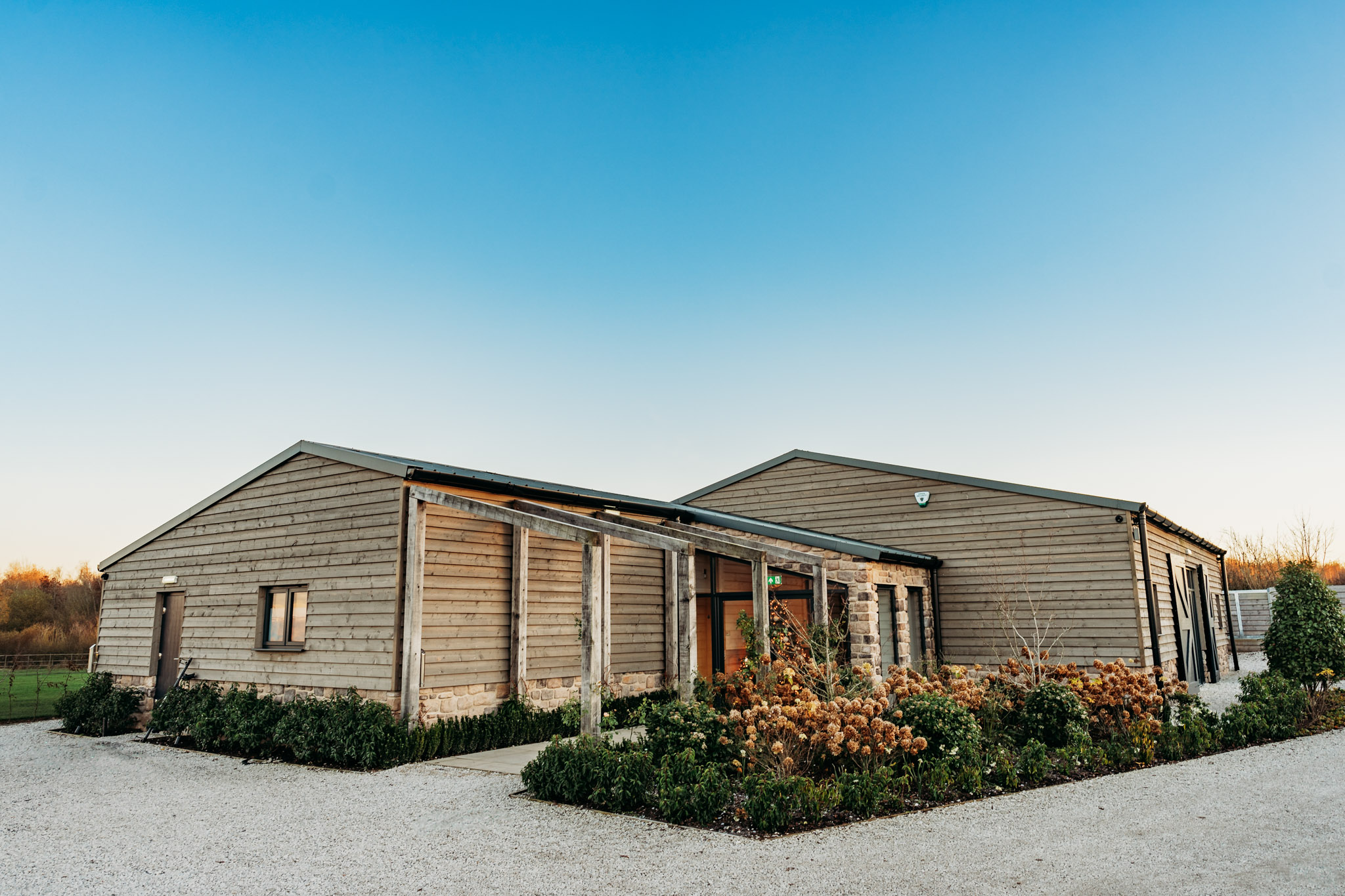Stretton Manor Barn - A modern barn-style building with wooden siding, surrounded by landscaped shrubs and gravel paths at Stretton Manor Barn, set against a clear blue sky and open fields.
